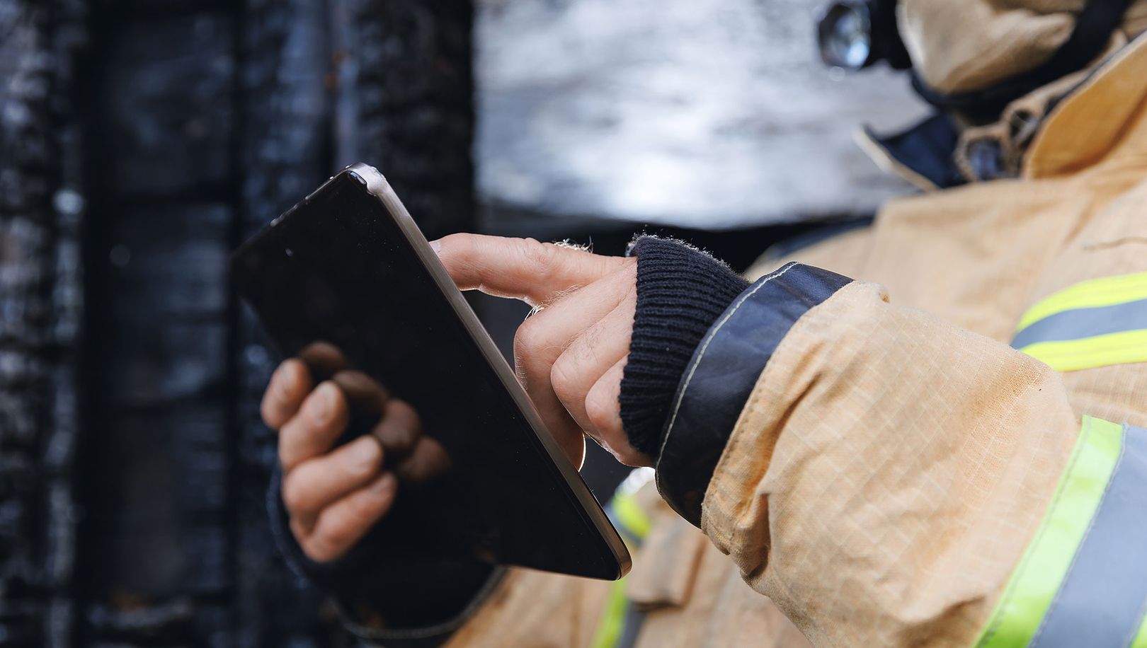 Person in Feuerwehrschutzausrüstung mit gelb-reflektierender Jacke bedient ein Tablet vor einem angerußten Hintergrund. Digitale Dokumentation im Brandschutzeinsatz