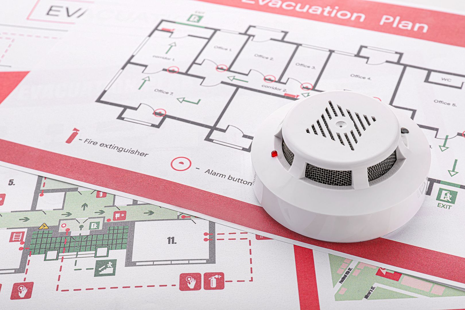 Weißer Rauchmelder mit roter LED liegt auf einem Evakuierungsplan mit eingezeichneten Fluchtwegen, Feuerlöscherstandorten und Alarmknöpfen