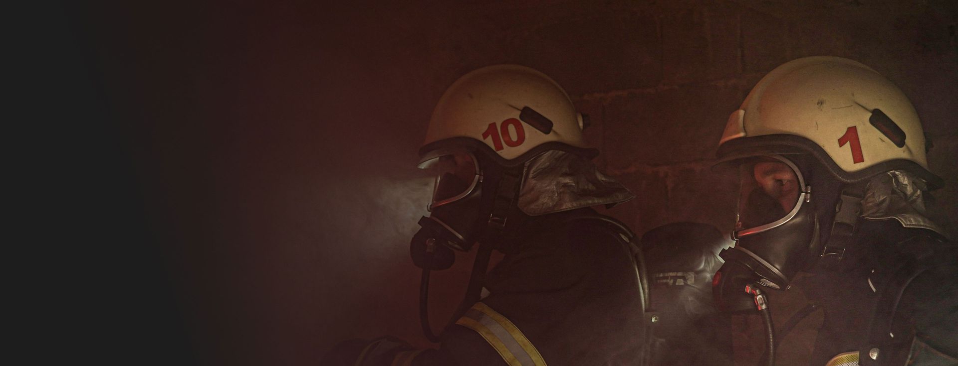 Zwei Feuerwehrleute in voller Schutzkleidung und mit Atemschutzmasken in einem verrauchten Raum. Einer der Feuerwehrleute trägt den Helm mit der Nummer 10. Dies veranschaulicht die Ernsthaftigkeit und Notwendigkeit des Brandschutzes.